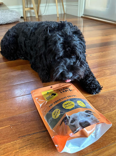 Black dog lying on a wooden floor next to a package of Lamb Liver dog treats from Bark with Buster.